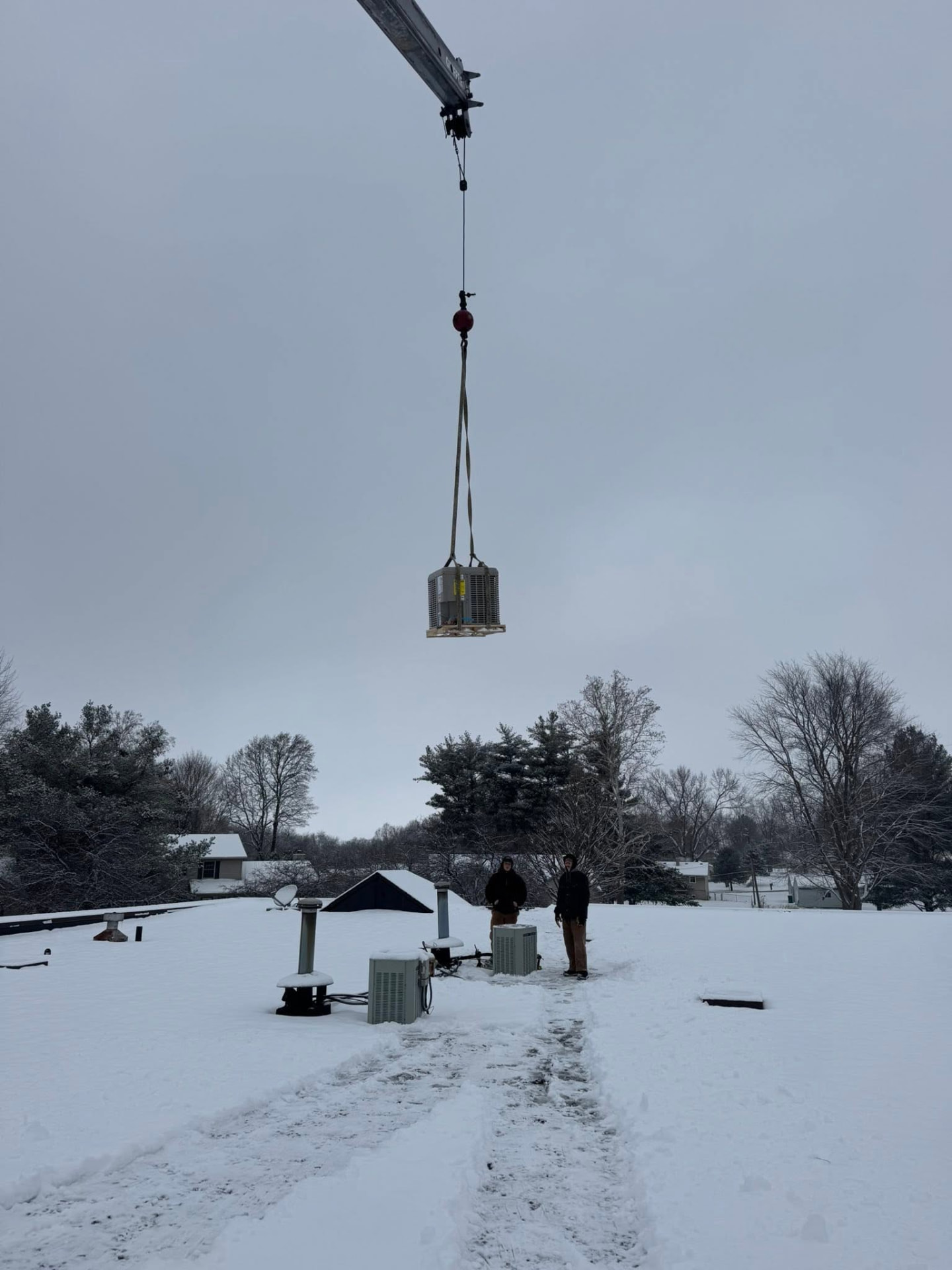 Crane lifting a rooftop condensing unit into place on a snowy winter install day